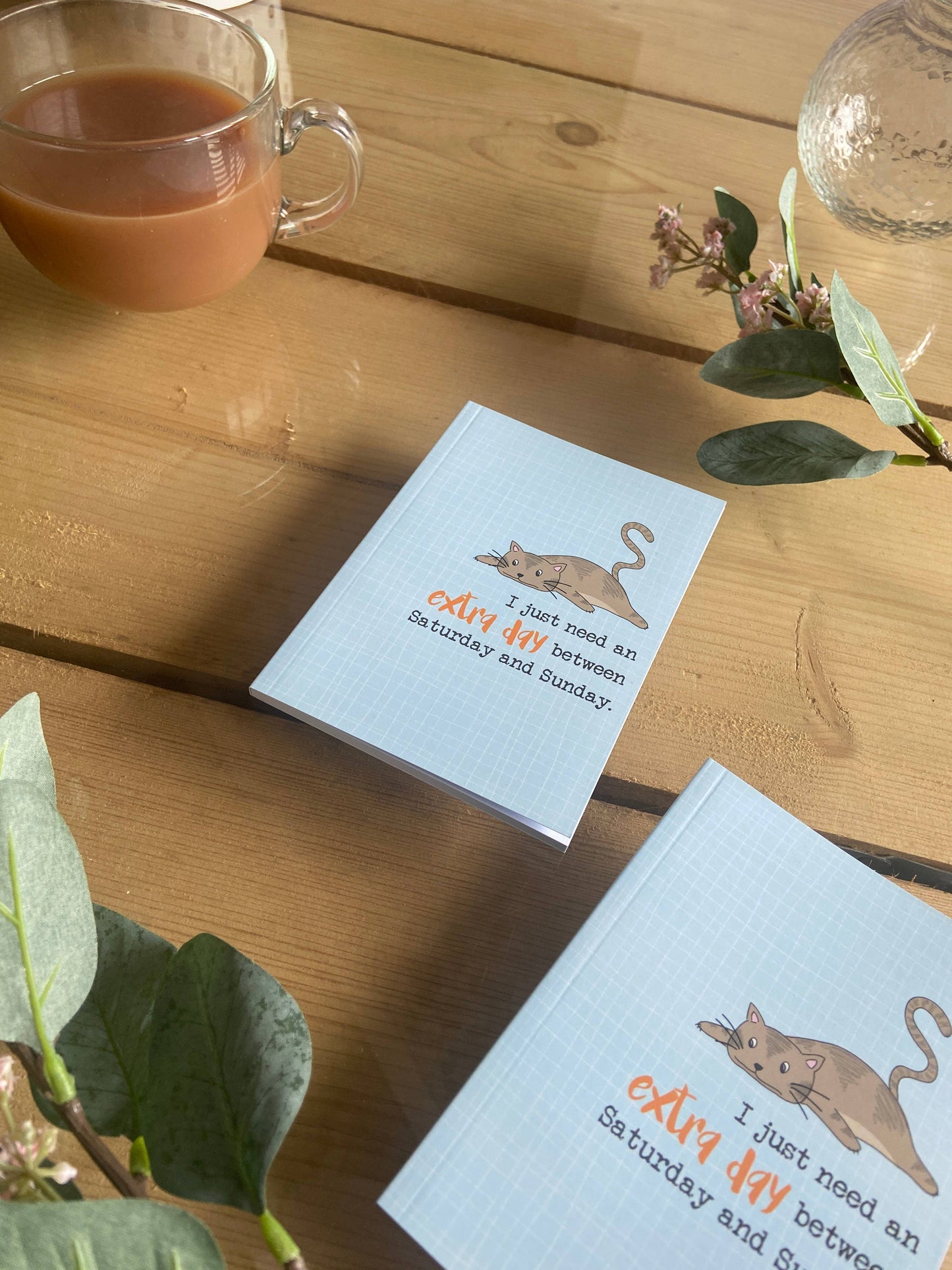 Two notebooks with cat illustrations and text on a wooden table with a cup of tea and flowers.