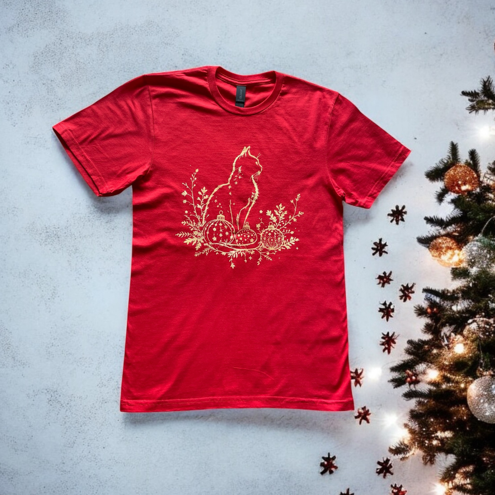 Red t-shirt with a gold cat design on a textured surface with a Christmas tree.