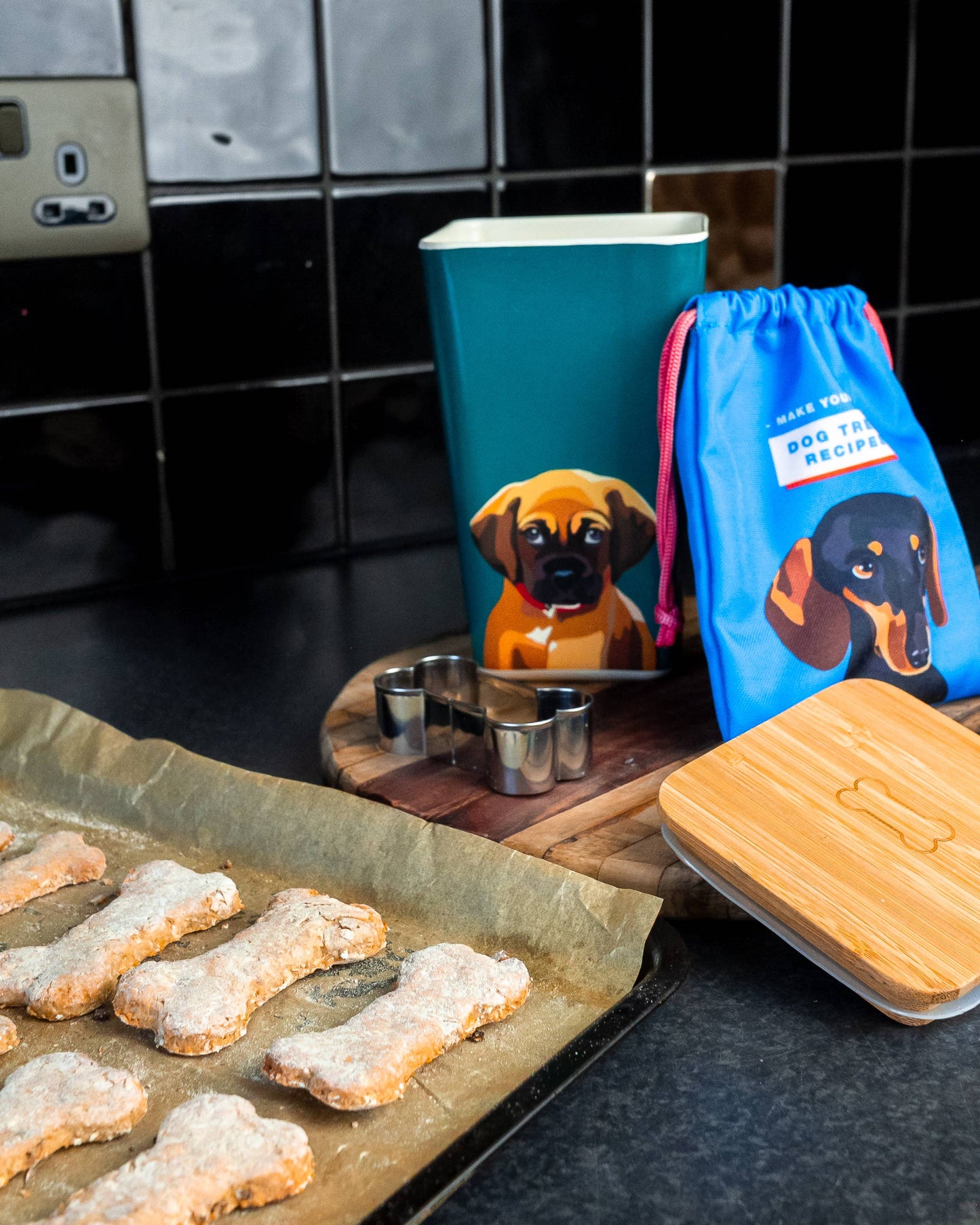 Dog treats on a baking tray with a mug, bowl, and blue bag featuring dog illustrations on a kitchen counter.