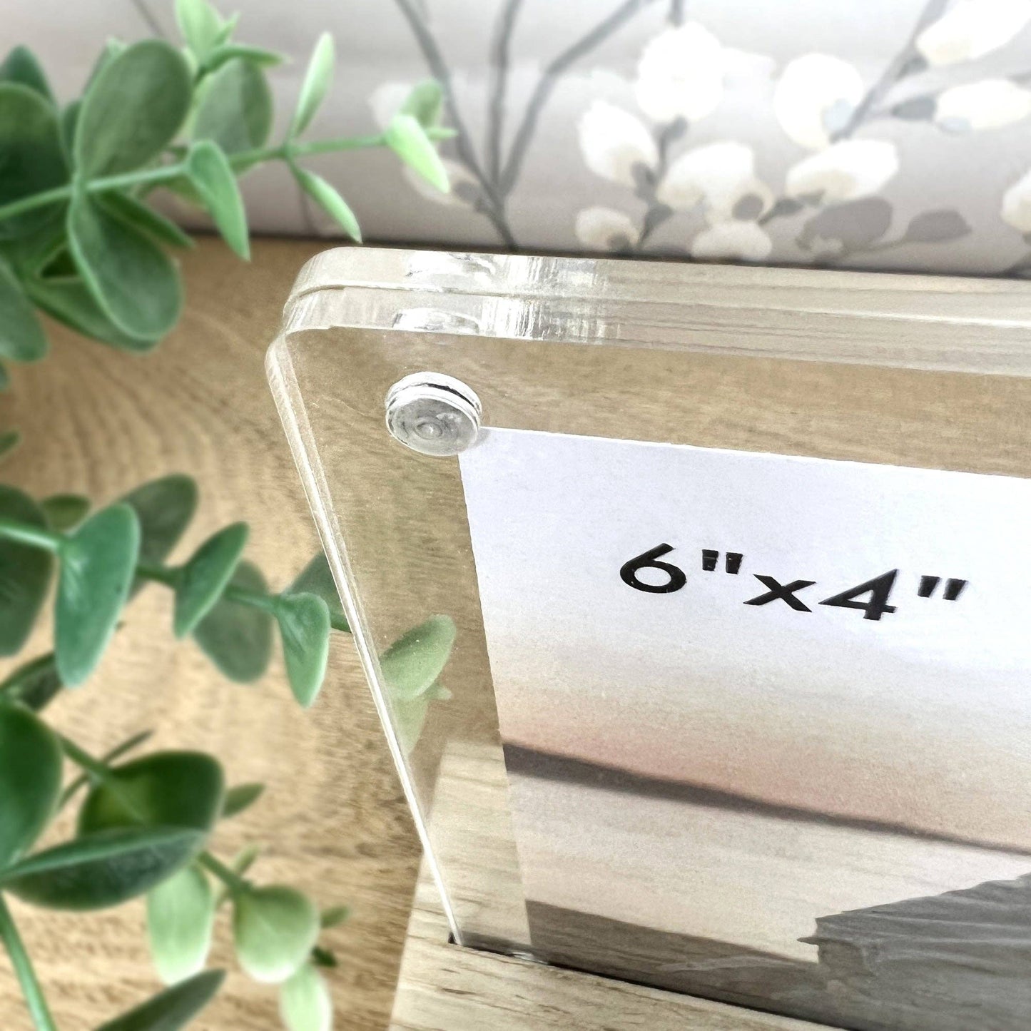 Clear acrylic photo frame with '6"x4"' label on a wooden surface with greenery.