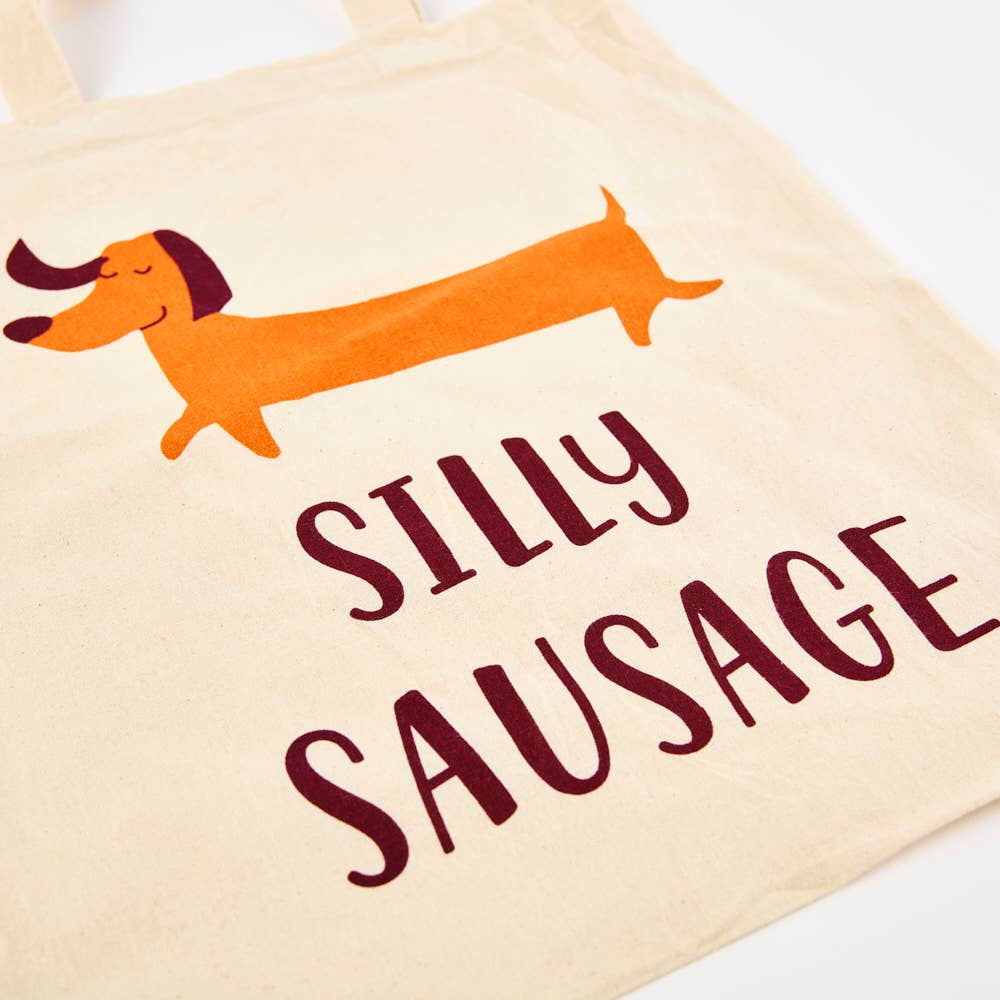 A white cotton tote bag with an illustration of a cartoon dog and the phrase 'SILLY SAUSAGE' printed on the front.