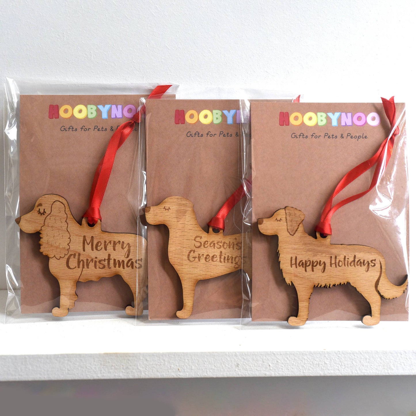 Three wooden dog-shaped Christmas ornaments with red ribbons and engraved text in clear packaging.