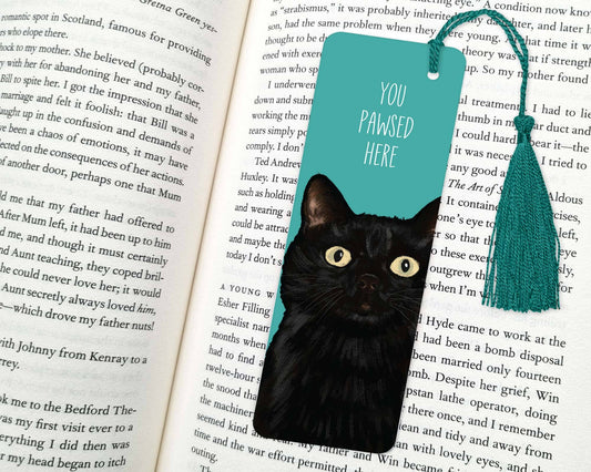 Black cat peeking out from behind a teal bookmark with 'You Paused Here' text on an open book.