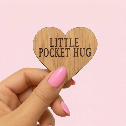 Wooden heart-shaped object with 'LITTLE POCKET HUG' text held by a hand with pink nails against a pink background