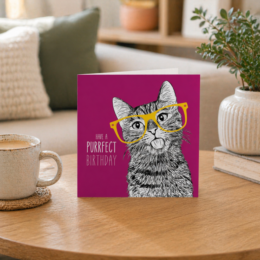 Card with a cat illustration and 'Purrfect Birthday' text on a table with a cup of coffee.