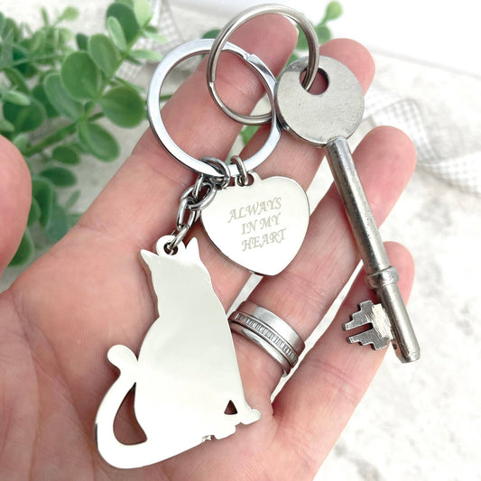 A stainless steel keyring with a silhouette cat shape and a heart-shaped charm that has 'Always in my heart' engraved on it, held in a hand with a key attached to it.