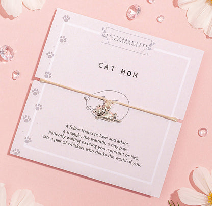 A silver-colored cat pendant on a nude-colored string bracelet, presented on a card with 'CAT MOM' text and a descriptive poem.