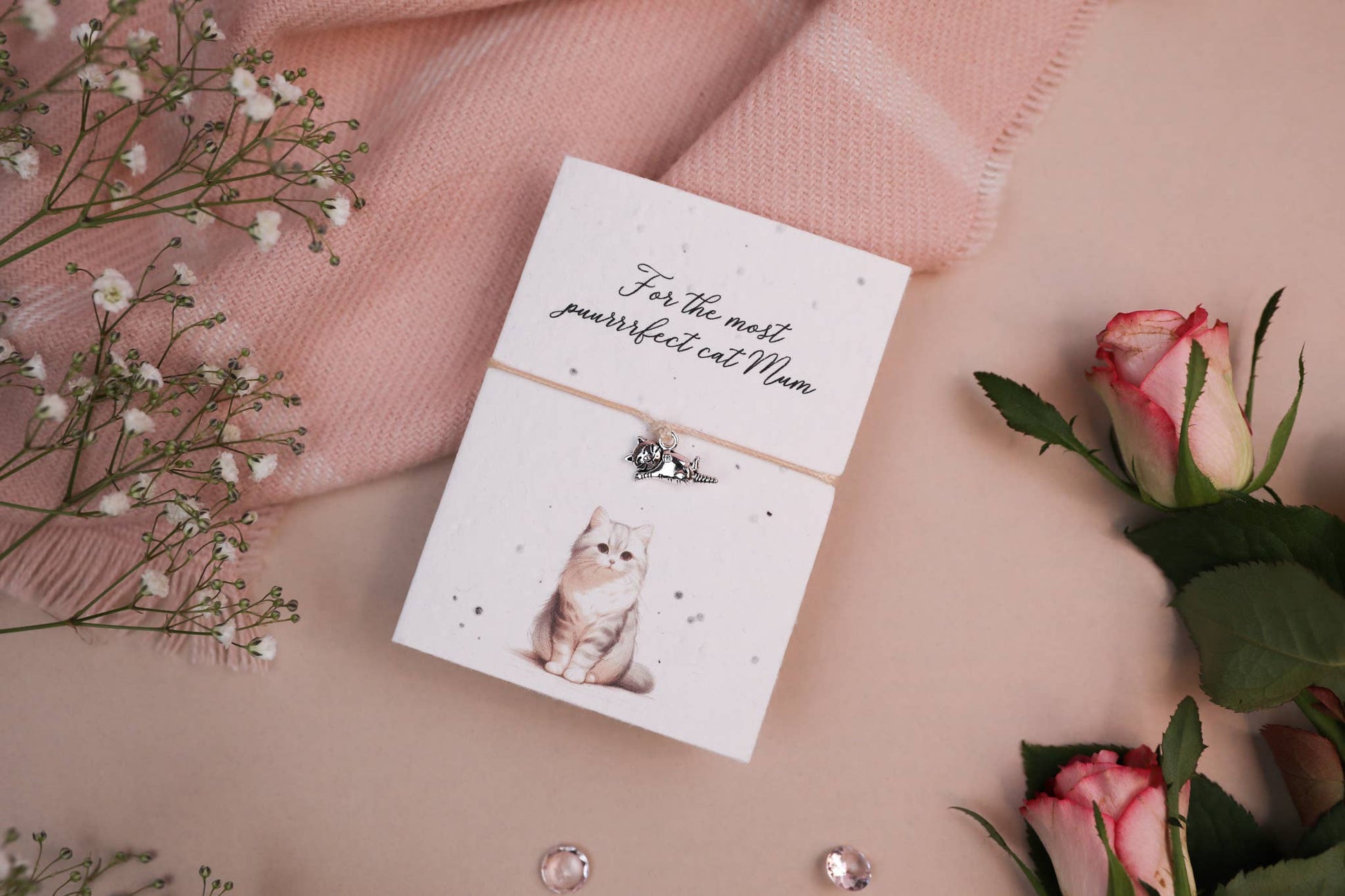 A set of floral-themed products including a card with a cat illustration and a wish bracelet, presented on a pink background with some decorative flowers and pearls.