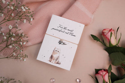 A set of floral-themed products including a card with a cat illustration and a wish bracelet, presented on a pink background with some decorative flowers and pearls.