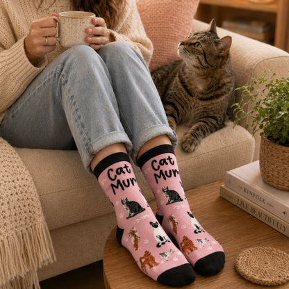 Person wearing pink socks with cat patterns sitting on a couch with a cat and a mug.