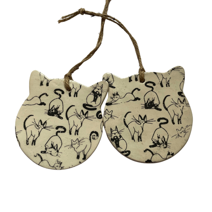 Two cat-shaped ceramic ornaments with black line drawings of cats on a beige background, hanging from a string.