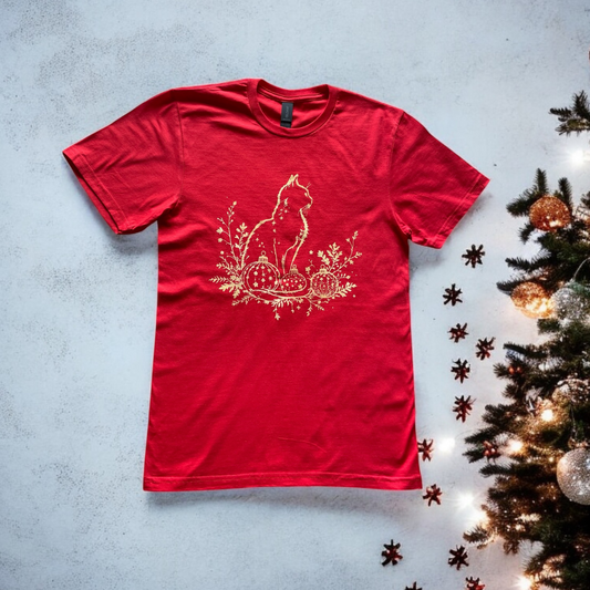 Red t-shirt with a gold cat design on a textured surface with a Christmas tree.