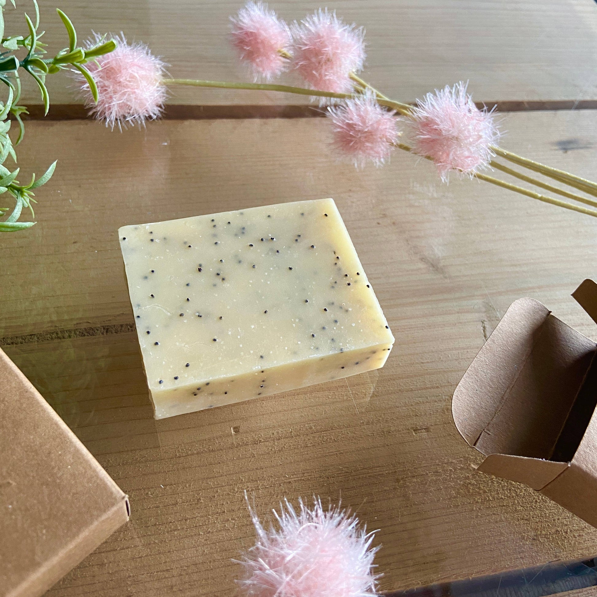 Bar of soap with black specks on a wooden surface with pink decorative flowers and cardboard boxes.