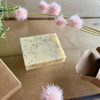 Bar of soap with black specks on a wooden surface with pink decorative flowers and cardboard boxes.