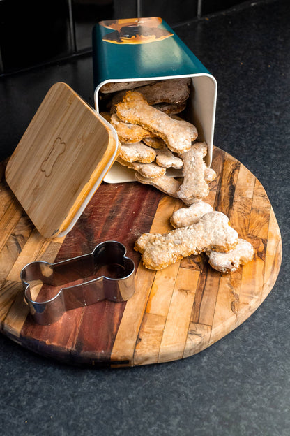 Dog treats on a wooden board with a cookie cutter and bamboo tin.
