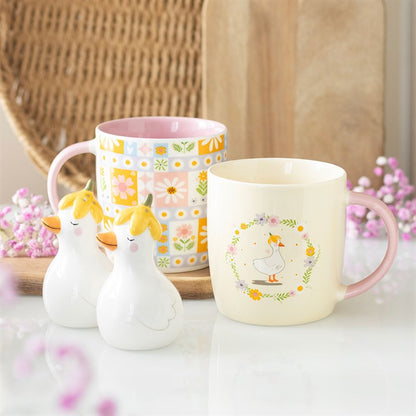 Two mugs with duck designs on a surface with flowers and a woven basket in the background.