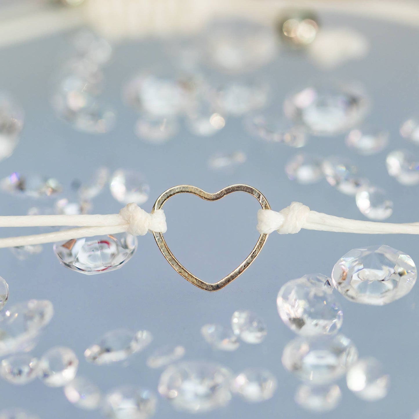 A cord bracelet with a stainless steel heart on a blue backdrop and clear diamonds scattered around. Close up shot. 