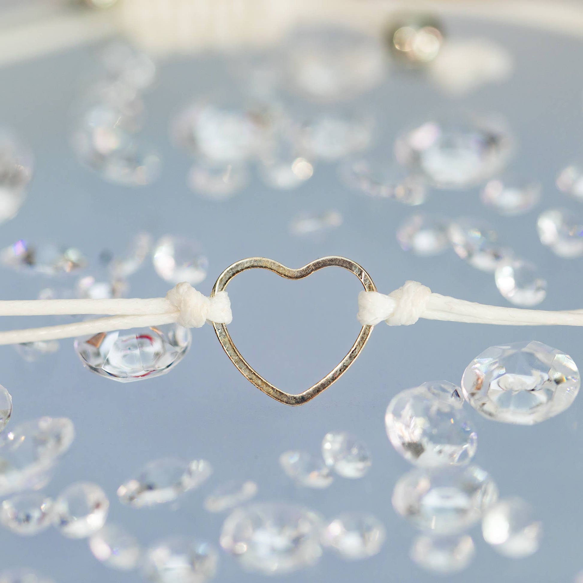 A cord bracelet with a stainless steel heart on a blue backdrop and clear diamonds scattered around. Close up shot. 