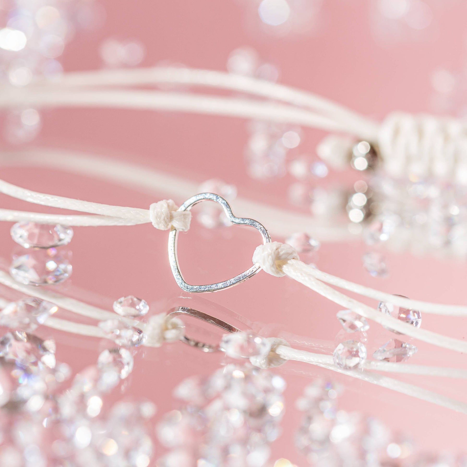 A cord bracelet with a stainless steel heart on a pink backdrop and clear diamonds scattered around. Close up shot.