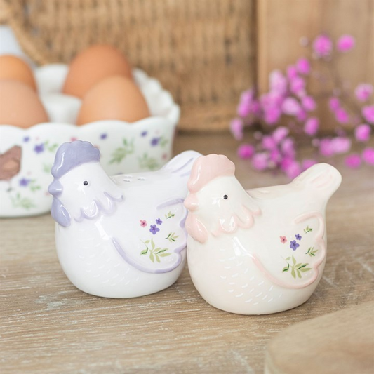 Two ceramic chicken figures, one purple and one pink, on a wooden surface with eggs and flowers in the background.