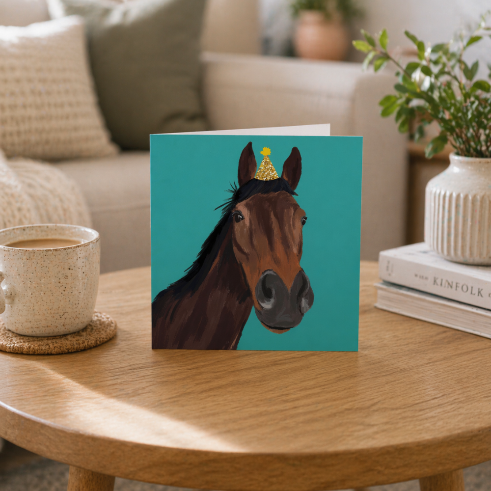 A teal birthday card with a horse on it wearing a party hat on a coffee table. 