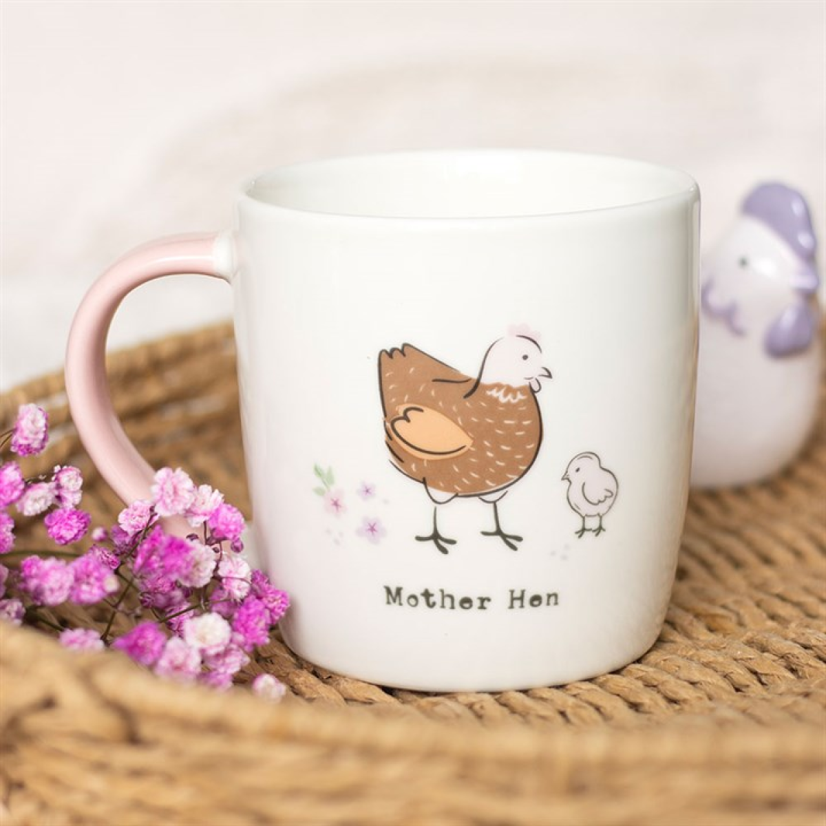 White mug with 'Mother Hen' design featuring a chicken and chick, placed on a woven surface with pink flowers.