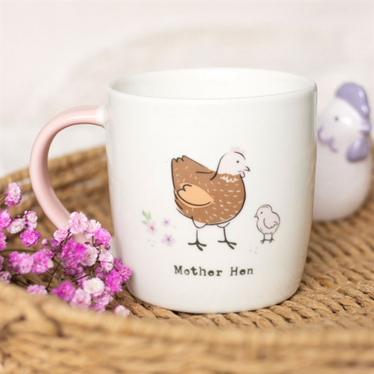 White mug with 'Mother Hen' design featuring a chicken and chick, placed on a woven surface with pink flowers.