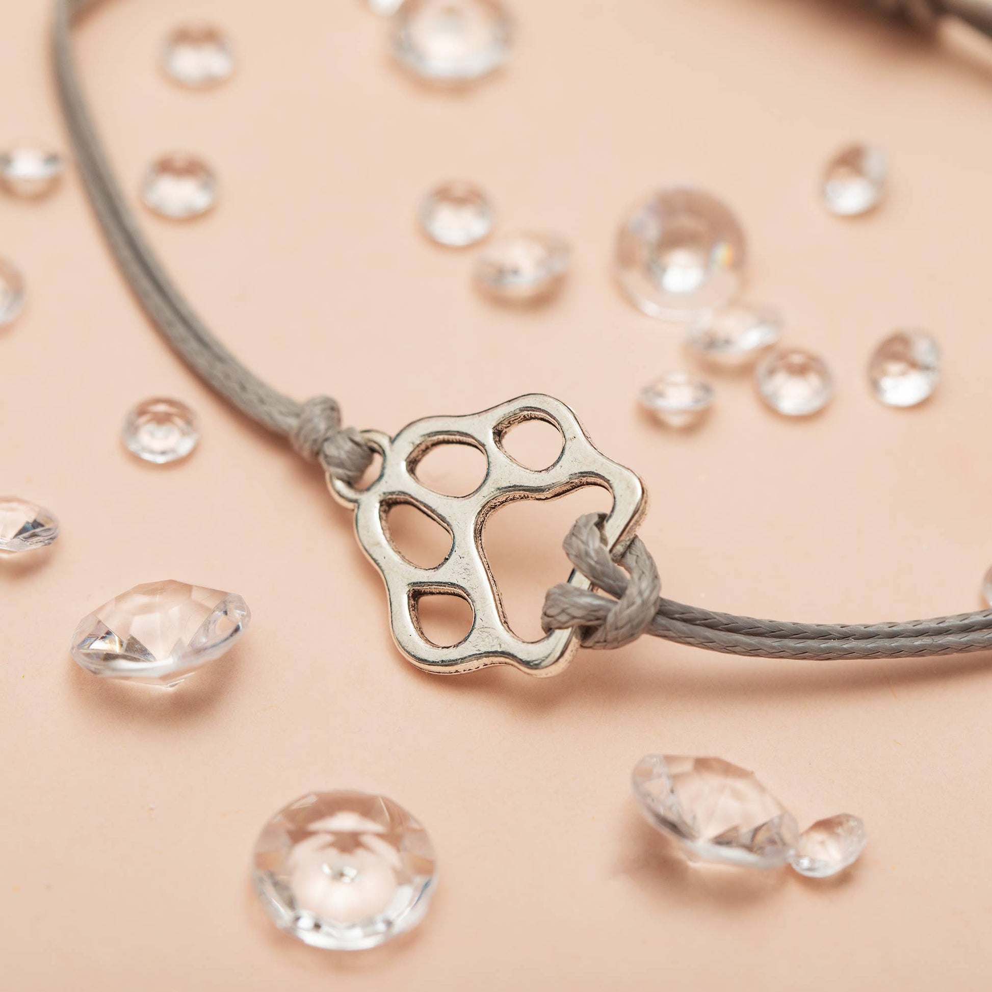 Close up shot of a grey bracelet with a dog paw charm on a peach backdrop and diamonds scattered around. 