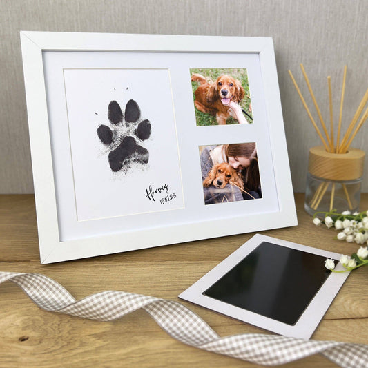 A paw print keepsake photo frame kit displayed on a wooden table with a white frame, black ink pad, and two small photos of a dog.