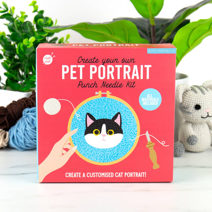 A punch needle kit for creating a cat portrait, displayed in front of a plant. The kit is packaged in a box with a picture of a cat and a person's hand using a punch needle.