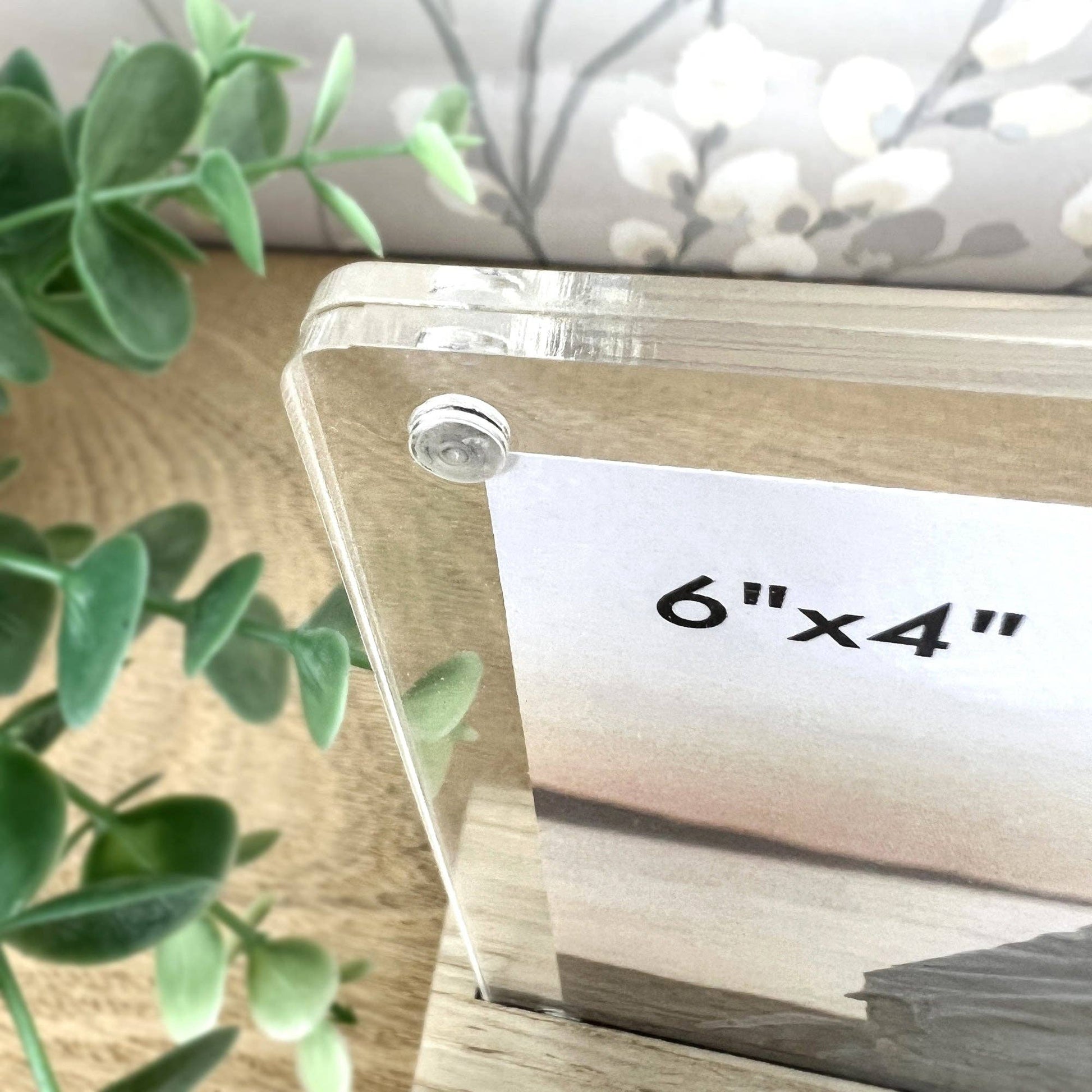 Clear acrylic photo frame with '6"x4"' label on a wooden surface with greenery.