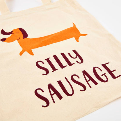 A white cotton tote bag with an illustration of a cartoon dog and the phrase 'SILLY SAUSAGE' printed on the front.