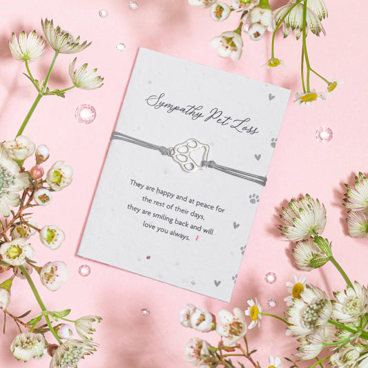 A sympathy card with a poem for pet loss placed next to a dainty bracelet made of zinc alloy and polyester thread, displayed on a pink background surrounded by white flowers.